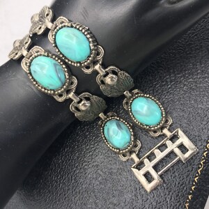 Op de afbeelding: Vintage zilverkleurige armband met ovale turquoise stenen met een gemarmerd effect. De armband heeft sierlijk metaalwerk met blad- en bloemaccenten en een sluiting. Een klassiek sieraad.