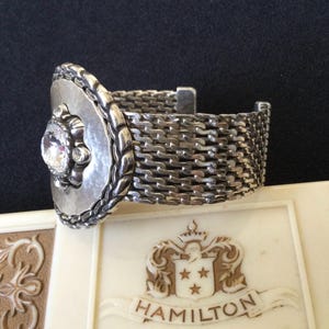 May include: A silver-toned bracelet with a woven band and a decorative front piece. The front piece features a large, clear gemstone surrounded by smaller stones and a textured, oval frame. The bracelet rests on a cream-colored surface.