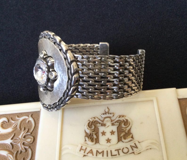 May include: A silver-toned bracelet with a woven band and a decorative front piece. The front piece features a large, clear gemstone surrounded by smaller stones and a textured, oval frame. The bracelet rests on a cream-colored surface.