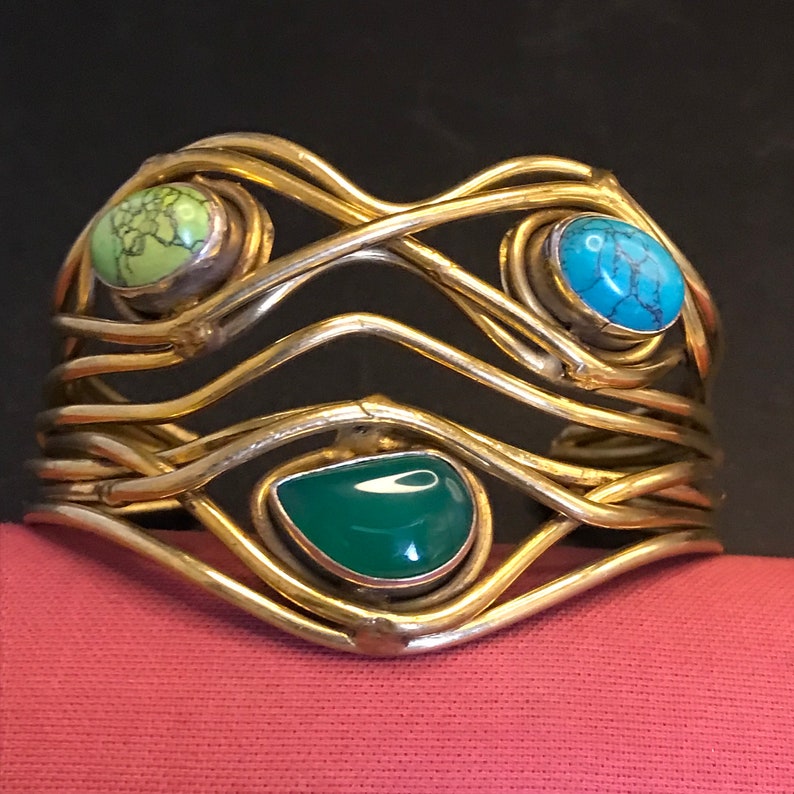 May include: A gold metal bracelet with a woven design and three colorful stones. The stones are green, turquoise, and a light green color.