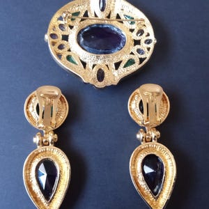 Peut inclure: Une broche et des boucles d'oreilles assorties dor&eacute;es avec une grande pierre violette fonc&eacute;e sertie dans un motif de filigrane. La broche a un fermoir &agrave; aiguille et les boucles d'oreilles ont des fermoirs &agrave; clip.