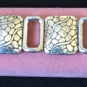May include: A silver-toned link bracelet featuring four square-shaped segments with a raised, textured pebble pattern alternating with three smooth, open rectangular links. The bracelet includes a metal clasp mechanism on the left side.
