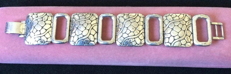 May include: A silver-toned link bracelet featuring four square-shaped segments with a raised, textured pebble pattern alternating with three smooth, open rectangular links. The bracelet includes a metal clasp mechanism on the left side.