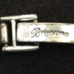 Może przedstawiać: Zbliżenie srebrnego metalowego zapięcia z wygrawerowanym słowem "Brightono". Zapięcie jest częścią większego elementu, prawdopodobnie biżuterii lub torebki. Metal ma lekko postarzany wygląd.