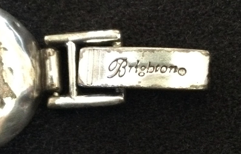 Może przedstawiać: Zbliżenie srebrnego metalowego zapięcia z wygrawerowanym słowem "Brightono". Zapięcie jest częścią większego elementu, prawdopodobnie biżuterii lub torebki. Metal ma lekko postarzany wygląd.