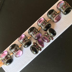 Peut inclure: Un bracelet vintage orn&eacute; de pierres pr&eacute;cieuses ovales altern&eacute;es noires, violettes et roses. Les pierres sont serties dans des montures en m&eacute;tal argent&eacute; orn&eacute;es de petits accents rouges, reli&eacute;es par des maillons dor&eacute;s.