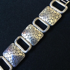May include: A silver-toned metal bracelet featuring a repeating pattern of square links with a textured, pebble-like surface alternating with smooth, open rectangular links. The design creates a geometric, link-style chain with a metallic finish.