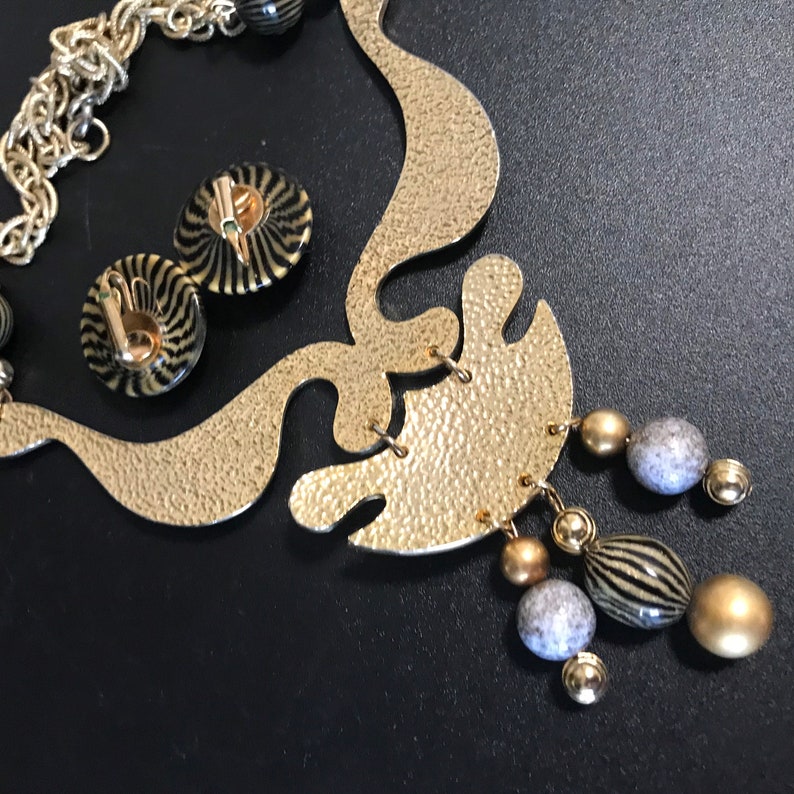 May include: Gold tone necklace with a large abstract pendant and black and white striped beads. The pendant has a textured surface and is attached to a chain with a lobster clasp. The necklace also includes a pair of matching earrings with black and white striped designs.