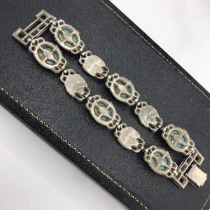 Op de afbeelding: Zilverkleurige armband met ovale en ruitvormige turkooizen stenen. De armband heeft een rechthoekige sluiting en wordt gepresenteerd op een zwarte ondergrond. Waarschijnlijk vintage of antiek.