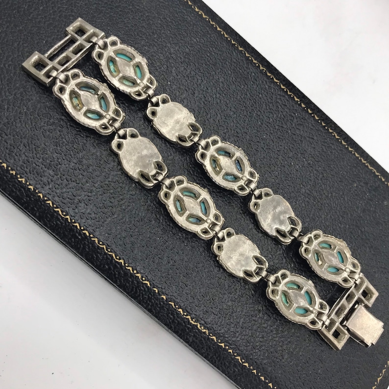 Op de afbeelding: Zilverkleurige armband met ovale en ruitvormige turkooizen stenen. De armband heeft een rechthoekige sluiting en wordt gepresenteerd op een zwarte ondergrond. Waarschijnlijk vintage of antiek.