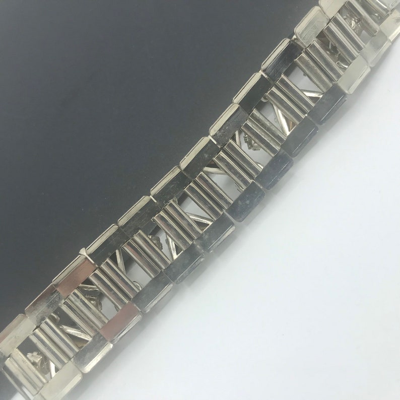 May include: A silver metal bracelet with a repeating pattern of rectangular links. The links are connected by a series of small, decorative pieces.