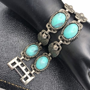 Op de afbeelding: Vintage zilverkleurige armband met ovale turquoise stenen met een marmer effect. De armband heeft sierlijk metaalwerk met blad- en bloemaccenten en een decoratieve sluiting. Een klassiek sieraad.