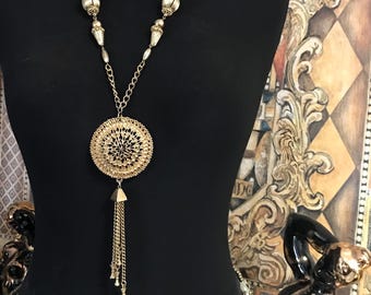 Peut inclure: Collier doré avec un grand pendentif orné. Le pendentif présente un motif ajouré détaillé avec un accent central sombre et une cascade de chaînes pendantes. Le collier est présenté sur un mannequin noir.