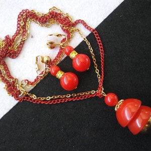 May include: A red and gold necklace and earrings set. The necklace has a layered chain with a red pendant made of three stacked round beads. The earrings are clip-on and feature a single red round bead.