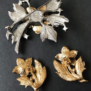 Puede incluir: Un conjunto de broche y pendientes florales plateados y dorados. El broche presenta un dise&ntilde;o detallado de flores y hojas. Los pendientes son dorados, con un dise&ntilde;o de flores y hojas. El conjunto es probablemente vintage.
