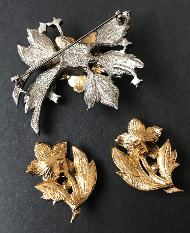 Puede incluir: Un conjunto de broche y pendientes florales plateados y dorados. El broche presenta un dise&ntilde;o detallado de flores y hojas. Los pendientes son dorados, con un dise&ntilde;o de flores y hojas. El conjunto es probablemente vintage.