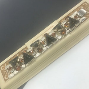 May include: A silver tone bracelet featuring a row of dark green and amber coloured triangular rhinestones. The bracelet is presented in a white box with a gold floral design.