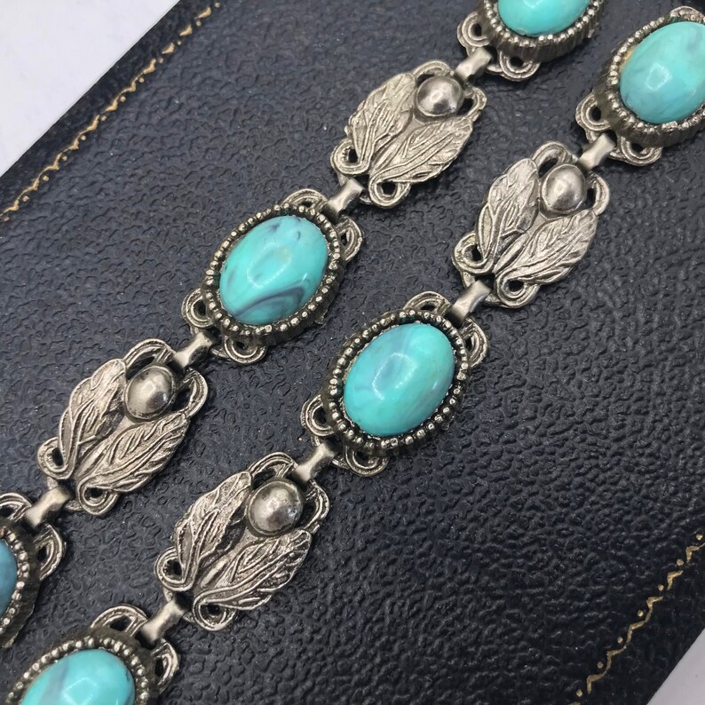 Op de afbeelding: Een vintage zilverkleurige armband met ovale turquoise stenen. De schakels van de armband zijn verbonden door decoratieve bladvormige elementen en kleine zilveren kralen. De turquoise stenen hebben een gemarmerd uiterlijk.