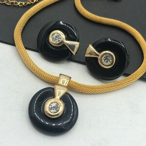 May include: A gold chain necklace with a black pendant featuring a round design with a clear rhinestone in the center. Matching earrings with black round designs and clear rhinestones are also included.