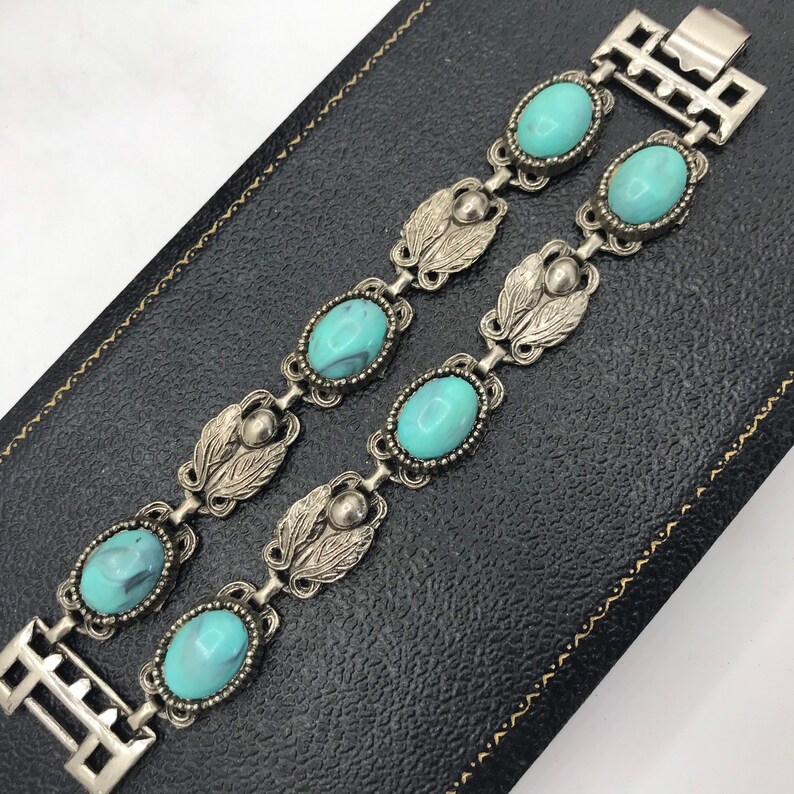 Op de afbeelding: Twee zilverkleurige armbanden met ovale, turquoise stenen en bladvormige accenten. De armbanden zijn verbonden door een rechthoekige sluiting. De sieraden worden gepresenteerd op een zwarte ondergrond.