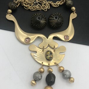 May include: A gold tone statement necklace with a geometric design and black and white striped beads. The necklace has a chain link and a large pendant with a gold tone finish. The pendant has a geometric design with a small iridescent stone in the center. The necklace also includes a pair of matching earrings with black and white striped beads.
