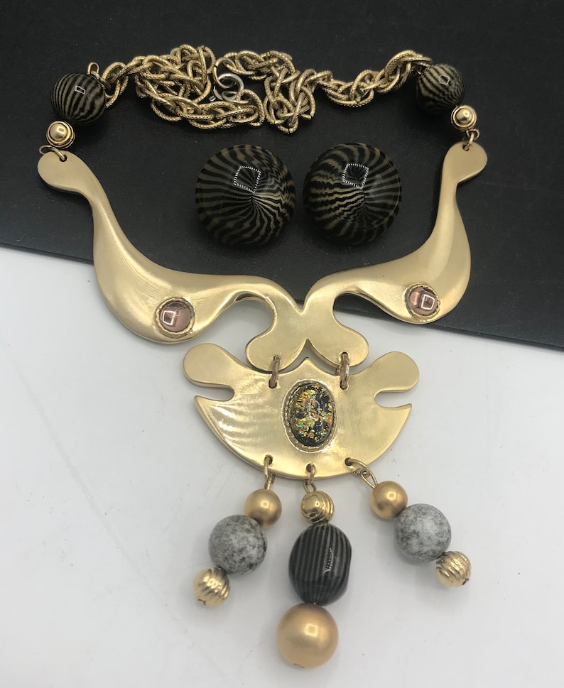 May include: A gold tone statement necklace with a geometric design and black and white striped beads. The necklace has a chain link and a large pendant with a gold tone finish. The pendant has a geometric design with a small iridescent stone in the center. The necklace also includes a pair of matching earrings with black and white striped beads.