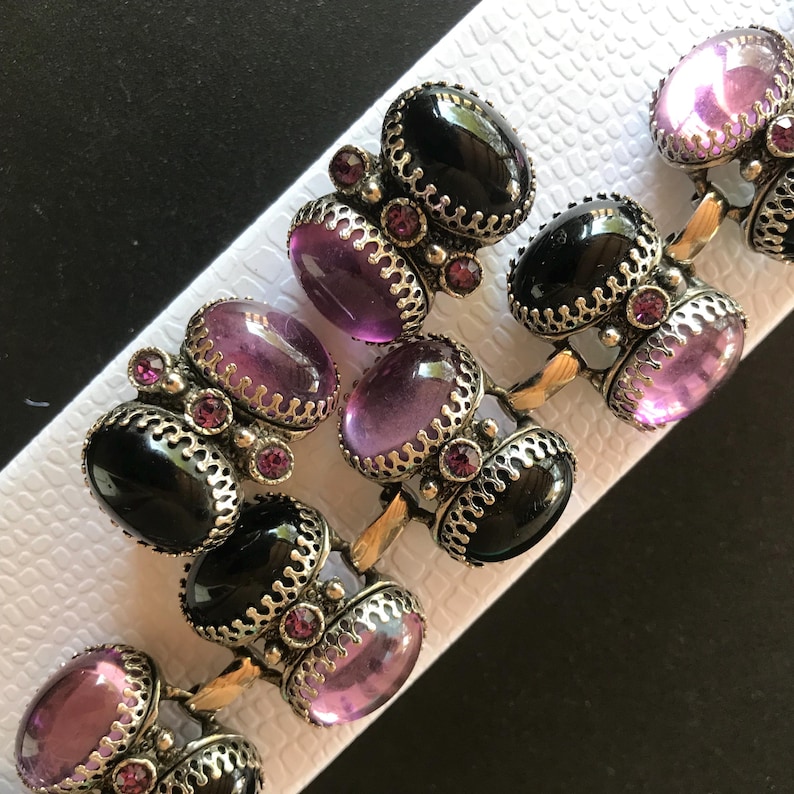 Peut inclure: Un bracelet vintage orn&eacute; de pierres ovales altern&eacute;es noires et lavande, serties dans un m&eacute;tal argent&eacute; orn&eacute;. De petites pierres roses rondes ornent le design. Le bracelet est pr&eacute;sent&eacute; sur une surface blanche textur&eacute;e.