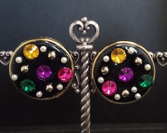Könnte beinhalten: Ein Paar schwarze und goldene Ohrringe mit einem runden Design. Die Ohrringe sind mit bunten Strasssteinen in den Farben Pink, Lila, Grün und Gelb verziert. Die Strasssteine sind in einem schwarzen Hintergrund mit einem goldenen Rand gefasst.