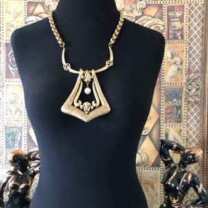 Può includere: Una collana dorata di tendenza con un grande ciondolo geometrico con superficie testurizzata e una singola goccia di perla. La collana è su un manichino nero.
