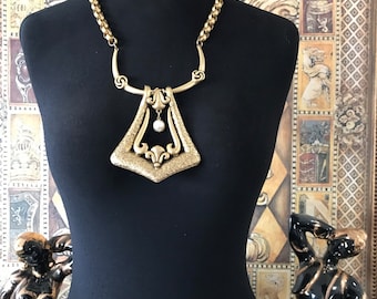 May include: A gold-tone statement necklace with a large, geometric pendant featuring a textured surface and a single pearl drop. The necklace is on a black mannequin.