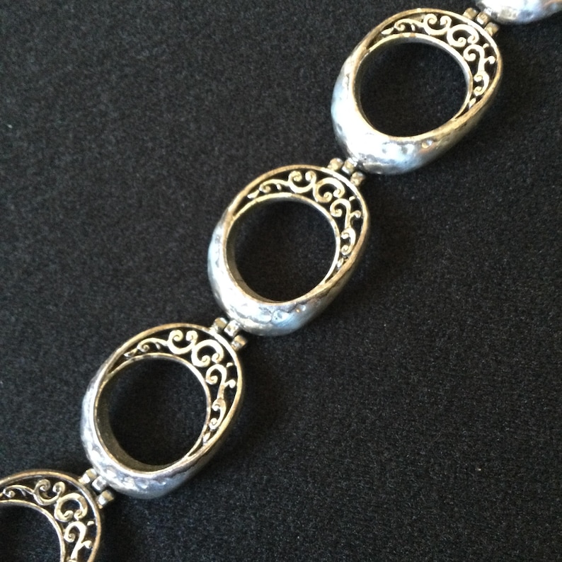 Może przedstawiać: Srebrna bransoletka z owalnymi ogniwami i skomplikowanymi ażurowymi wzorami filigranowymi. Bransoletka składa się z serii połączonych owalnych ogniw, z kt&oacute;rych każde ma szczeg&oacute;łowy, wirujący wz&oacute;r w owalnej ramie. Metal ma lekko młotkowaną fakturę.