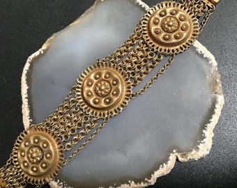 Peut inclure: Un bracelet en or avec plusieurs chaînes et des médaillons ronds décoratifs. Les médaillons ont un design en relief texturé avec un cercle central et des cercles plus petits autour.