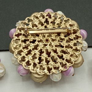 May include: A set of vintage jewelry featuring a large, gold-toned brooch with a filigree design and a cluster of pink and white beads. The set also includes two matching earrings with a similar design.