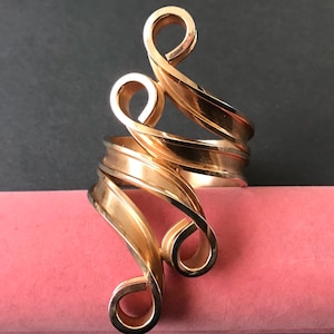 May include: A gold-toned metal ring with an open design. The ring features a series of curved, ribbon-like elements that wrap around the finger. The design includes multiple loops and swirls, creating a sculptural effect. The ring is displayed on a pink surface.