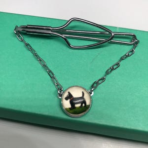 May include: A silver tie clip with a black and white image of a dog on a white background. The dog is wearing a red collar.