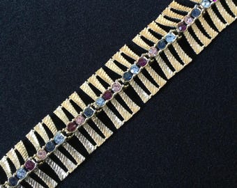 May include: Gold-toned bracelet with a textured, openwork design. A row of colorful, round gemstones in shades of blue, purple, and pink runs down the center. The bracelet is set against a dark background, highlighting its intricate details.