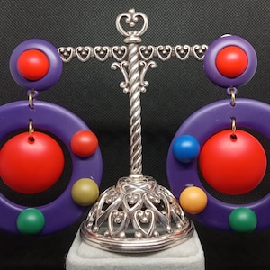 May include: A pair of purple and red statement earrings with a circle design and colorful accents. The earrings are displayed on a silver stand.