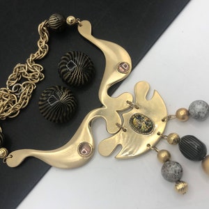 May include: A gold necklace with a large abstract pendant and black and white patterned beads. The pendant has a small, clear stone in the center. The necklace is accented with gray and gold beads.