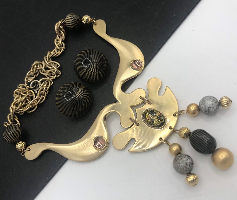 May include: A gold necklace with a large abstract pendant and black and white patterned beads. The pendant has a small, clear stone in the center. The necklace is accented with gray and gold beads.