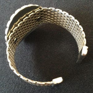 May include: Silver-toned cuff bracelet with a woven mesh design. The bracelet has a rectangular shape with a scalloped edge and a hinged opening. The metal has a shiny finish, reflecting light.
