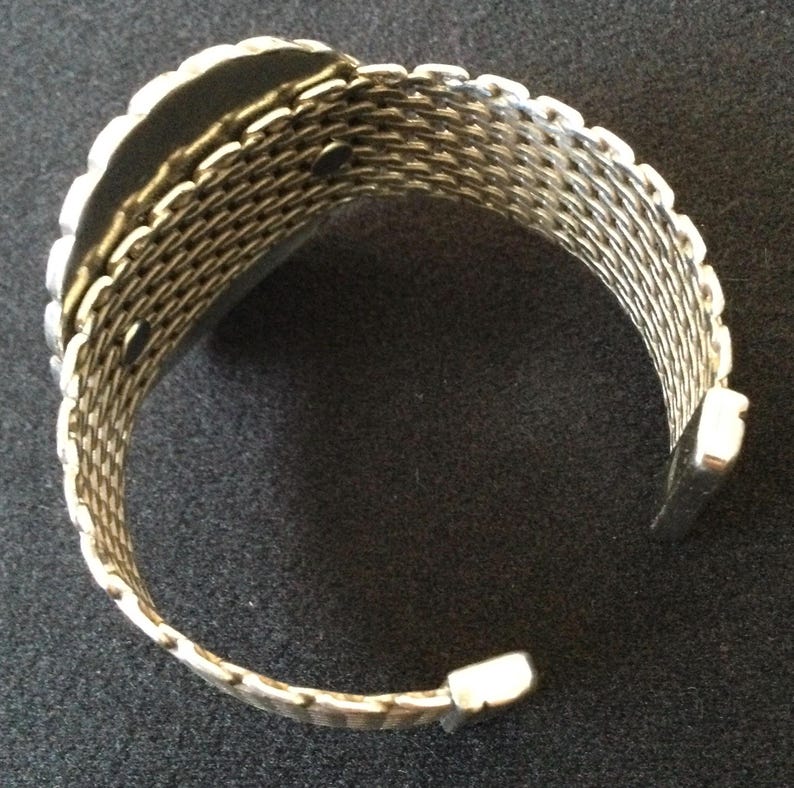 May include: Silver-toned cuff bracelet with a woven mesh design. The bracelet has a rectangular shape with a scalloped edge and a hinged opening. The metal has a shiny finish, reflecting light.
