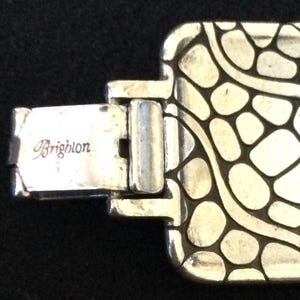 May include: A silver-toned metal bracelet featuring a rectangular link with a raised, irregular pebble-like pattern and dark recessed lines. The clasp mechanism is clearly visible and engraved with the brand name Brighton in script lettering.