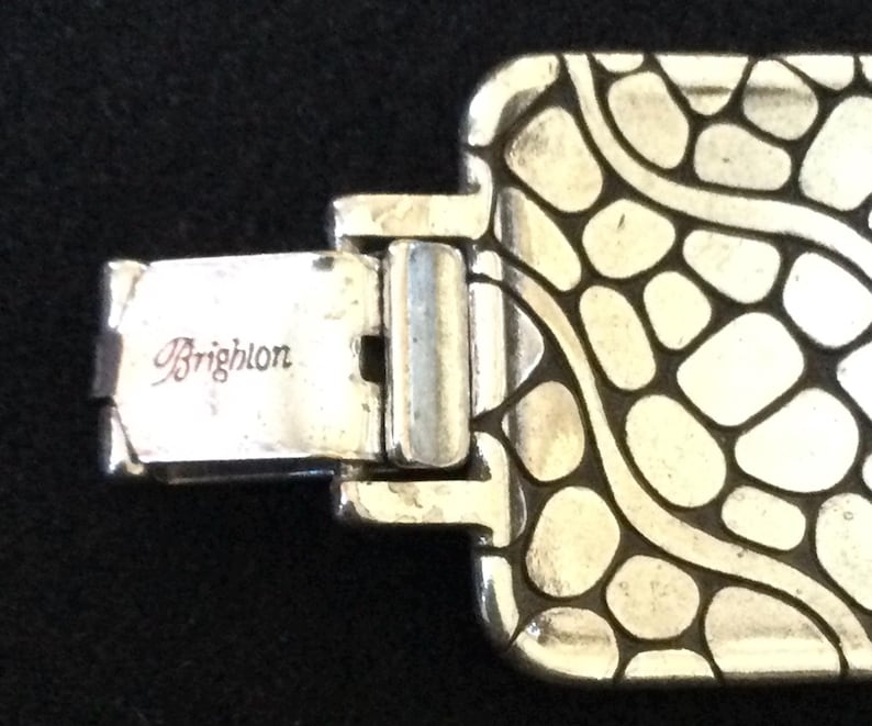 May include: A silver-toned metal bracelet featuring a rectangular link with a raised, irregular pebble-like pattern and dark recessed lines. The clasp mechanism is clearly visible and engraved with the brand name Brighton in script lettering.