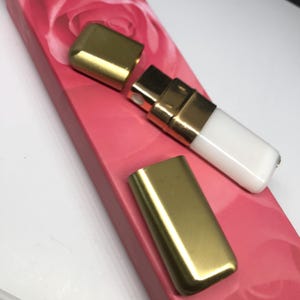 May include: A gold and white refillable perfume atomizer with a gold cap. The atomizer is sitting on a pink surface with a rose design.
