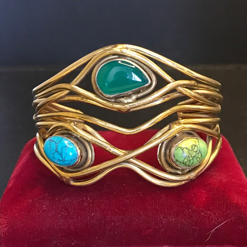 May include: A gold-toned metal bracelet with a woven design and three colorful stones. The stones are green, turquoise, and a light green with brown markings.