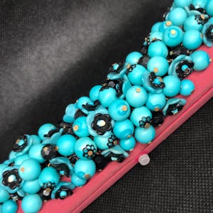 May include: A close-up of a turquoise and black beaded necklace with gold accents. The beads are arranged in a cluster and are attached to a pink fabric backing.