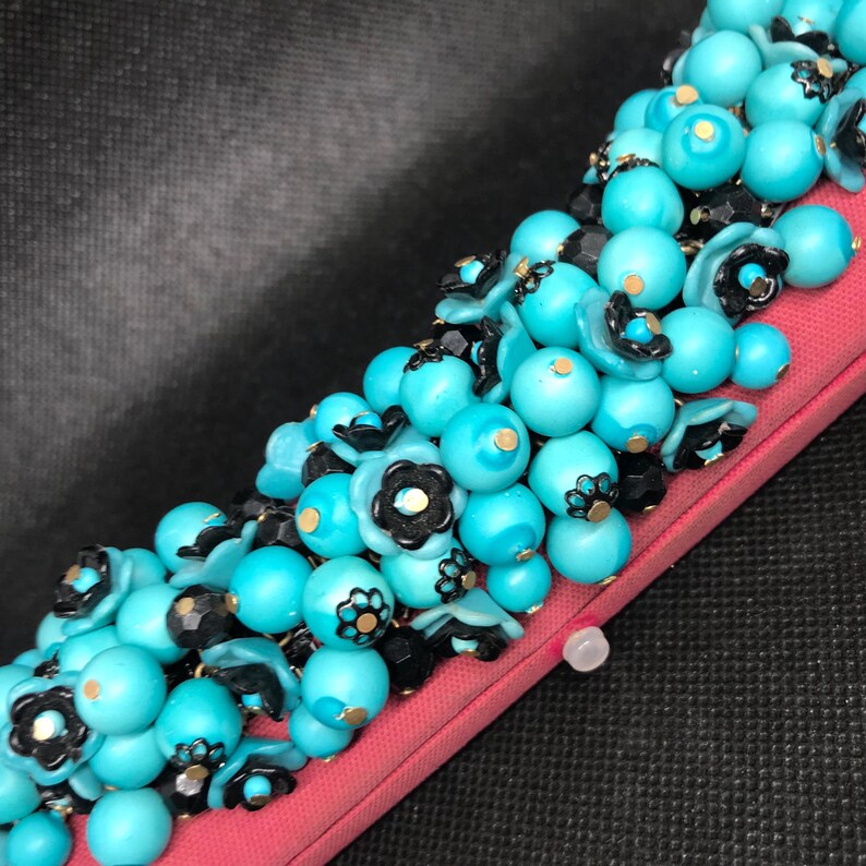 May include: A close-up of a turquoise and black beaded necklace with gold accents. The beads are arranged in a cluster and are attached to a pink fabric backing.