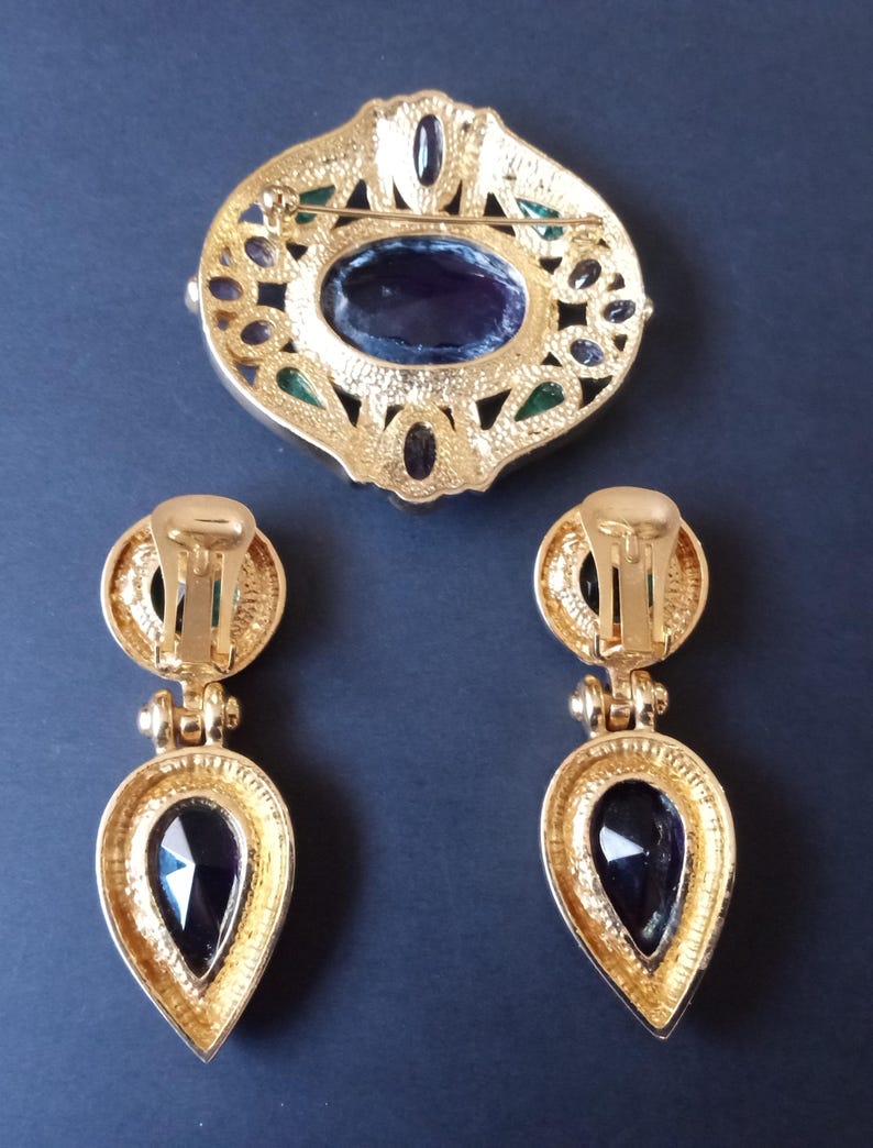 Peut inclure: Une broche et des boucles d'oreilles assorties dor&eacute;es avec une grande pierre violette fonc&eacute;e sertie dans un motif de filigrane. La broche a un fermoir &agrave; aiguille et les boucles d'oreilles ont des fermoirs &agrave; clip.