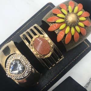 Puede incluir: Tres pulseras doradas con diferentes dise&ntilde;os. Una pulsera presenta una gran flor naranja y amarilla con un centro dorado. Otra pulsera tiene una piedra marr&oacute;n engastada en un marco dorado. La tercera pulsera tiene un coraz&oacute;n de cristal transparente rodeado de cristales m&aacute;s peque&ntilde;os.