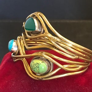 May include: A gold-toned bangle bracelet with three different colored stones. The stones are green, turquoise, and a light green color. The bracelet is made of multiple strands of wire that are woven together.